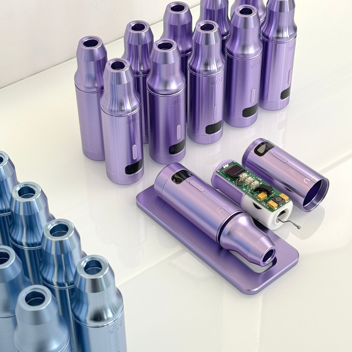 Carcasas moradas y azules de máquinas de maquillaje permanente sobre una superficie blanca, con el interior de una de ellas mostrando la electrónica.